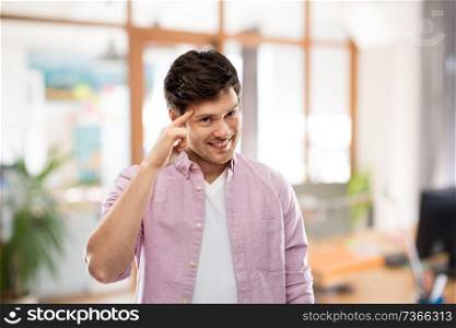 idea, intelligence and imagination concept - smiling young man pointing finger to his temple over office room background. smiling man pointing finger to his head at office
