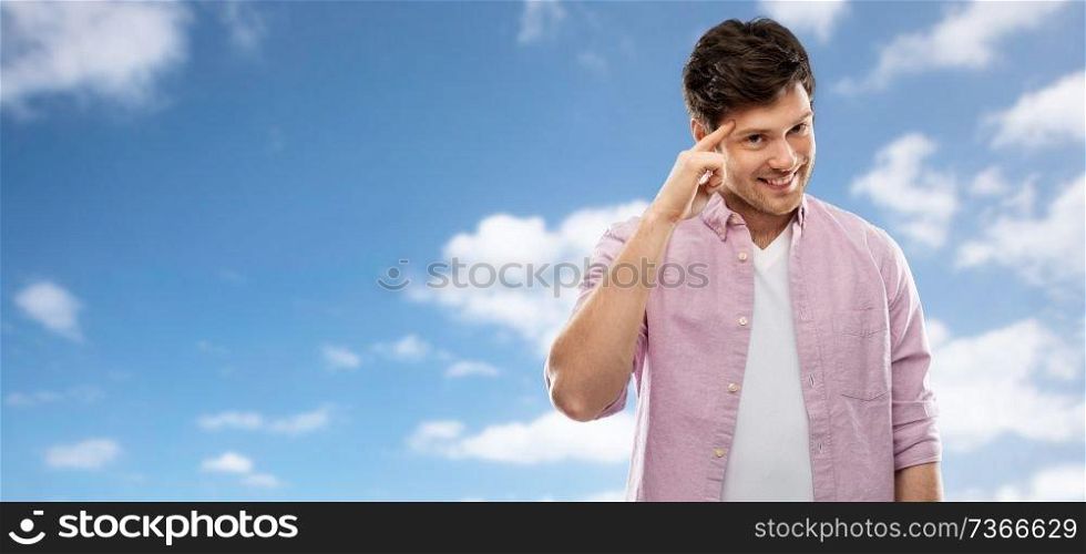 idea, intelligence and imagination concept - smiling young man pointing finger to his temple over blue sky and clouds background. smiling young man pointing finger to his head
