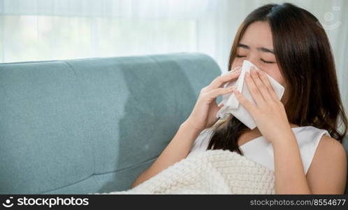 Ill Asian young woman cold covered with blanket sitting on sofa and sneeze with tissue paper at home, Female sick allergic blowing nose sneezing in tissue at home, Flu health care