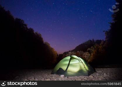 Illuminated green tent at night in the forest under dark blue sky with many stars