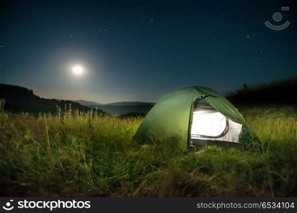 Illuminated green tent in the mountains at night under dark sky with many stars. Green tent in the mountains at night