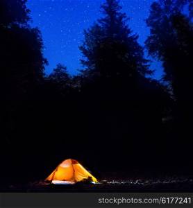 Illuminated orange tent at night in the forest under dark blue sky with many stars
