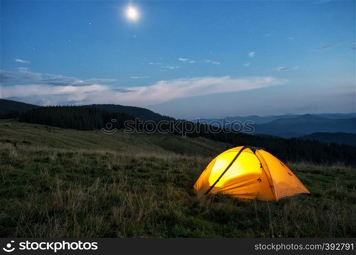 Illuminated orange tent in the mountains at dusk. The tent is lit from the inside. In the sky, the moon and the stars. Illuminated orange tent in mountains at dusk