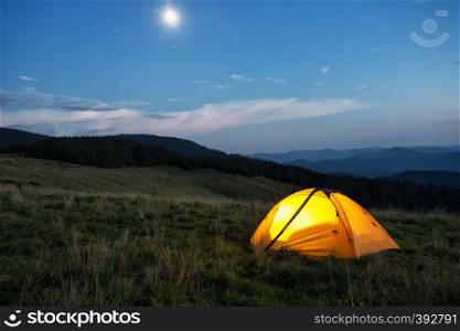 Illuminated orange tent in the mountains at dusk. The tent is lit from the inside. In the sky, the moon and the stars. Illuminated orange tent in mountains at dusk