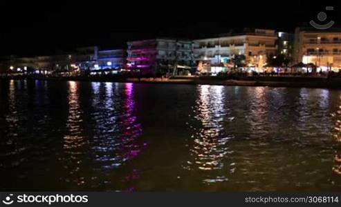 Illuminated waterfront buildings at night casting colourful reflections over a calm ocean