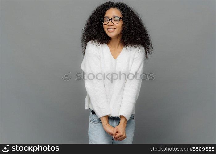 Ima≥of happy dreamy woman keeps hands to≥ther, has eyes shut, smi≤s positively, has frizzy hair, wears spectac≤s, white jumper and jeans, isolated over grey background, Good emotions concept