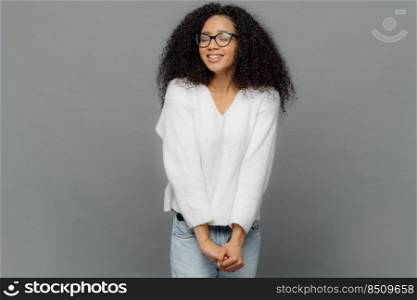 Ima≥of happy dreamy woman keeps hands to≥ther, has eyes shut, smi≤s positively, has frizzy hair, wears spectac≤s, white jumper and jeans, isolated over grey background, Good emotions concept