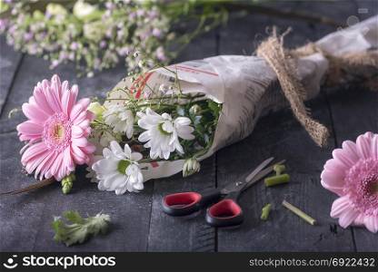 Image about the activity of wrapping flowers with a bouquet of chrysanthemum packed in newspaper, surrounded by blooms and scissors, on a rustic wooden background.