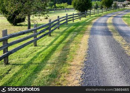 Image of a gravel road in the countryside with a split rail fence, field and woods. Layout in horizontal format.