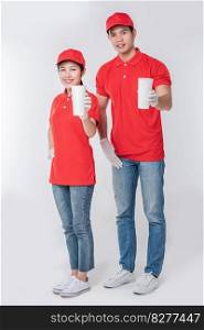 Image of a happy young delivery man in red cap blank t-shirt uniform standing with empty white paper cup isolated on light gray background studio
