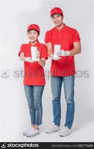 Image of a happy young delivery man in red cap blank t-shirt uniform standing with empty white paper cup isolated on light gray background studio