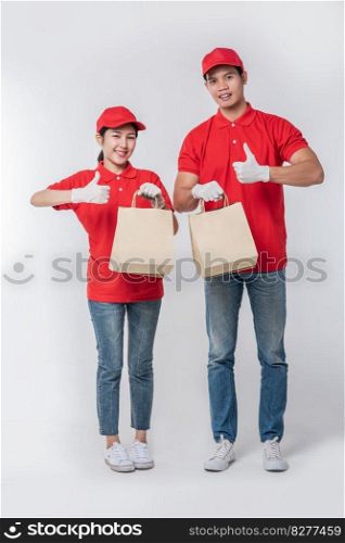 Image of a happy young delivery man in red cap blank t-shirt uniform standing with empty brown craft paper packet isolated on light gray background studio