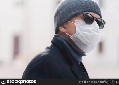 Image of adult man wears shades and protective medical mask, walks outdoor during coronavirus epidemic, looks aside, thinks how to overcome virus, blurred background. Male has seasonal allergy