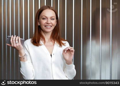 Image of attractive brown haired young female in white clothes, works in company, holds cell phone, sends text messages, checks email box, makes money transaction, expresses positive emotions