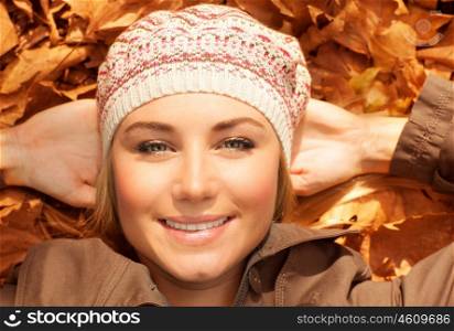 Image of attractive female laying down on the ground in autumn park, happy blond girl in fashionable beret resting on golden fall foliage, good mood, warm sunny day, autumnal woods, fun concept