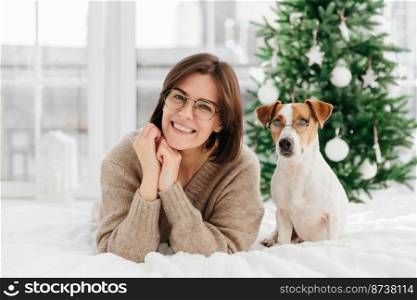 Image of attractive woman with gentle smile, has dark hair, wears transparent glasses spends time with lovely pet, lie on white bed against New Year tree. Merry Christmas and happy winter holidays
