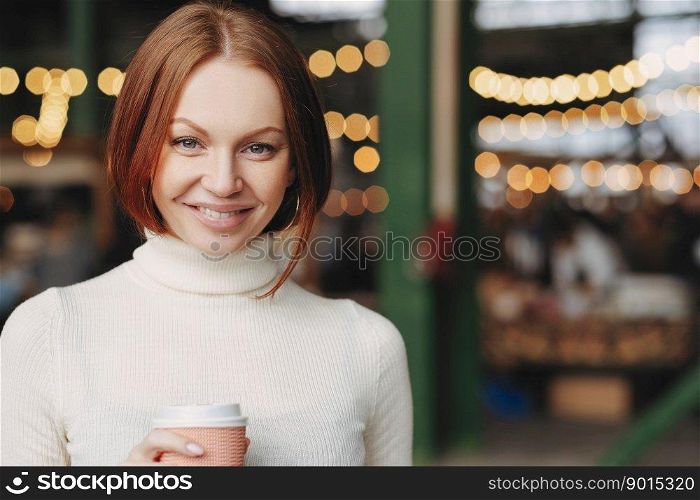 Image of attractive young woman with brown hair, gentle smile, wears white turtleneck sweater, holds paper cup of coffee, has pleased expression, poses over blurred background with copy space