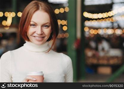 Image of attractive young woman with brown hair, gentle smile, wears white turtleneck sweater, holds paper cup of coffee, has pleased expression, poses over blurred background with copy space