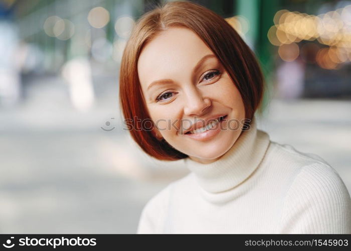 Image of attractive young woman with glad expression, looks directly at camera, has pleased look, wears turtleneck sweater, poses over blurred background, walks outside, expresses good emotions