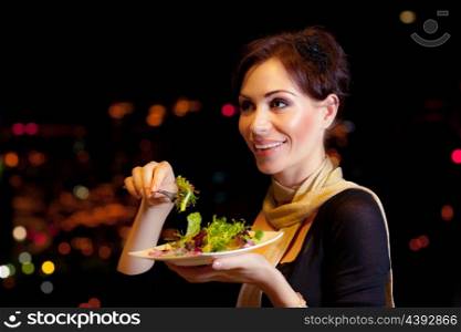 Image of beautiful female eating salad on the party, night lifestyle, attractive woman have dinner in restaurant outdoor, girl wearing dress holding in hand plate with meal, holiday celebration