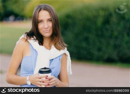 Image of beautiful young Caucasian woman with dark hair, wears casual clothes, holds disposable cup of coffee, stands agaist green nature background, has walk in park, looks directly at camera