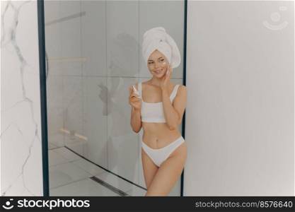 Image of beautiful young girl with towel on head holding moisturizing face cream while standing in bathroom after morning hygiene routine, isolated over light wall background. Beauty concept. Image of beautiful young girl with towel on head holding moisturizing face cream