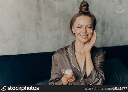 Image of caucasian cheerful woman in silk pajama smiling while gently applying moisturizing face cream isolated over concrete wall background in bedroom. Beauty and skincare concept. Image of caucasian cheerful woman in silk pajamas sitting on bed