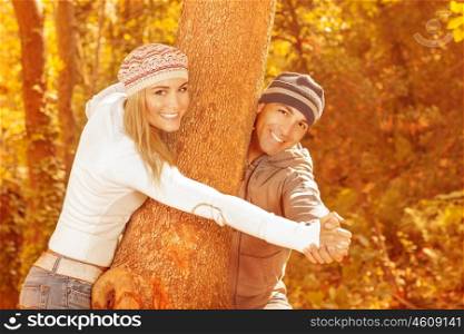 Image of cheerful couple playing around tree in autumnal park, happy young family having fun in autumn garden, sweet teenagers dating in fall woods, romantic relationship, love concept&#xA;