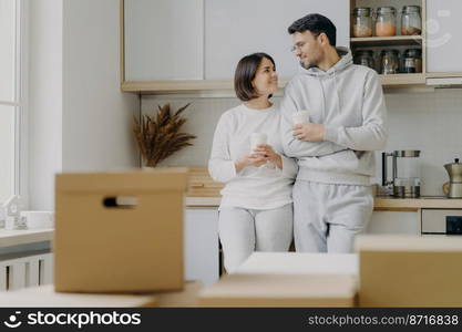 Image of cheerful woman and man have pleasant talk during coffee break, wear casual clothes, move in new apartment, pose with modern furnished kitchen, unpack cardboard boxes with household stuff