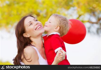 Image of cute little boy kissing mom, beautiful brunette woman with adorable child having fun outdoor in spring time, small kid with red balloon enjoying summer holiday, happy family, love concept