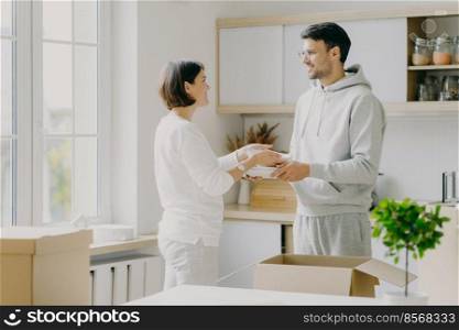 Image of delighted family couple unpack utensils with carton boxes, move in new home, pose against kitchen interior, looks happily at each other, busy unpacking different home stuff. Householding