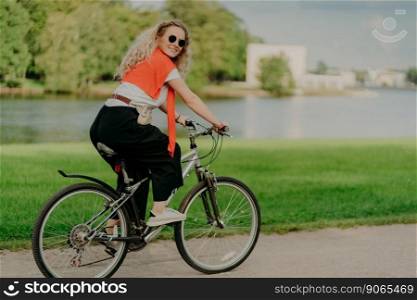 Image of female model rides bicycle, looks aside with cheerful expression, wears sunglasses, breathes fresh air, poses near lake and green trees, covers long distance, finds out something new