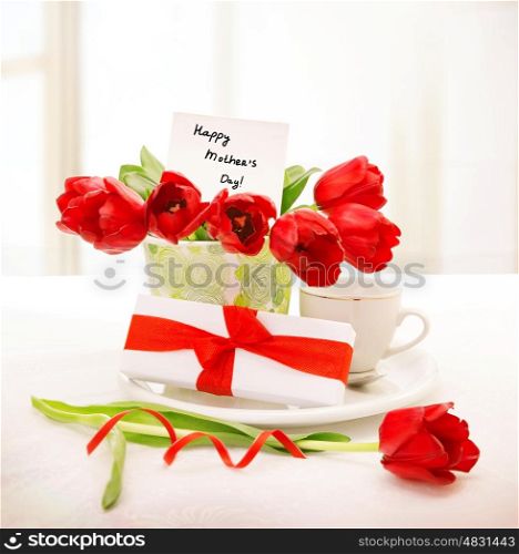 Image of fresh red tulips bouquet in beautiful vase with greeting postcard on the table, little white giftbox with ribbon, cup with morning coffee, breakfast for mommy, happy mothers day concept