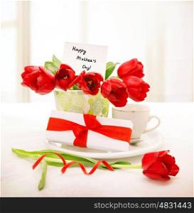 Image of fresh red tulips bouquet in beautiful vase with greeting postcard on the table, little white giftbox with ribbon, cup with morning coffee, breakfast for mommy, happy mothers day concept