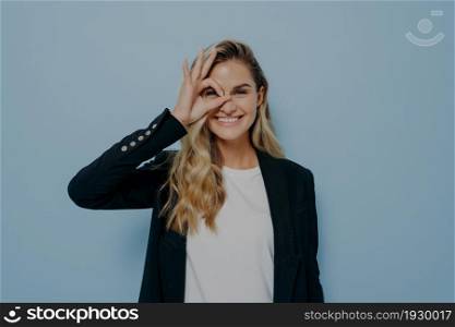 Image of glad happy young female with long blonde hair in black blazer and white shirt, making ok gesture with hand and looking through her fingers, rejoicing good day, showing how great she is doing. Image of glad happy young female with long blonde hair in black blazer making ok gesture with hand