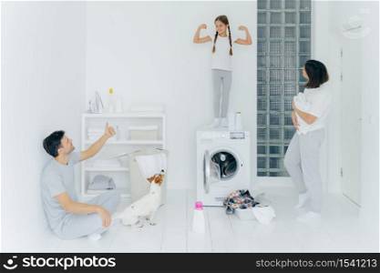Image of glad small child raises arms, shows biceps and stength, father shows like sign with thumb up, stand in washing room with pile of clothes in basin near washer, detergent. Well done work