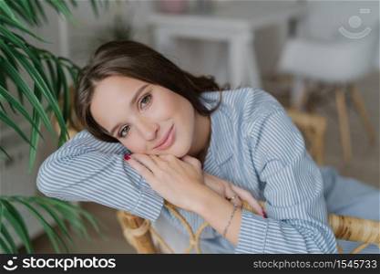 Image of good looking young female model with dark hair, sits on wooden chair, has relaxed facial expression, has charming smile, enjoys domestic calm atmosphere. Beauty and emotions concept