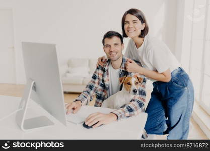 Image of happy brunette smiling woman leans at husbands shoulders who keyboards on computer, surf internet and search new apartment to buy on website, pose in spacious room with domestic dog