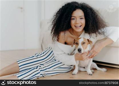 Image of happy curly woman plays with funny little dog, pose on floor in spacious room, sofa near, smiles broadly, embraces pet with love, dressed in fashionable clothes, has fun with animal