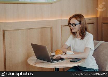 Image of happy smiling woman in earphones using laptop while sitting at cafe, young female freelancer working remotely on laptop computer in coffee shop, studying online during morning breakfast. Happy smiling woman in earphones using laptop while sitting at cafe