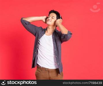 Image of happy young asian student man dress in casual shirt singing and listening to music with cell phone and wireless earphones isolated over red wall and copy space.