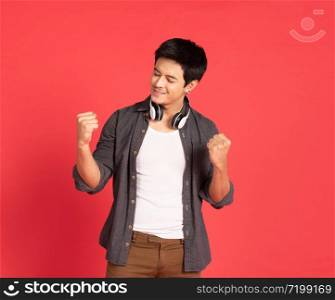 Image of happy young asian student man dress in casual shirt singing and listening to music with cell phone and wireless earphones isolated over red wall and copy space.