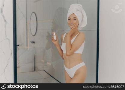 Image of happy young female with head wrapped in towel after having morning shower enjoying beauty care procedures while holding cosmetic lotion and applying some cream on face, smiling at camera. Happy young female with head wrapped in towel after enjoying daily beauty morning routine