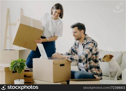 Image of happy young man keyboards something on laptop computer, has glad expression, sits on comfortable sofa with pet, surrounded with cardboard boxes, make online shopping, buy new furniture