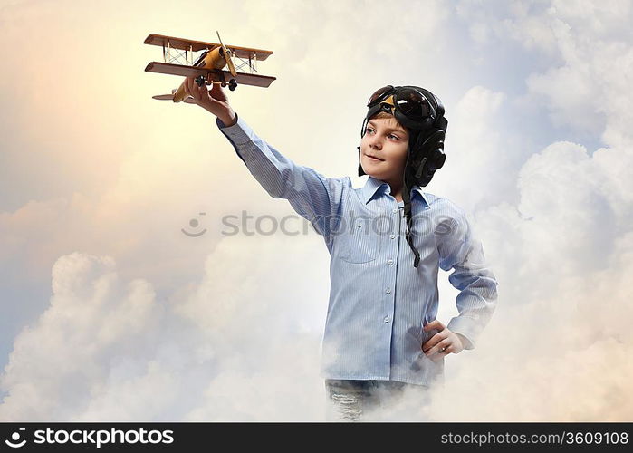 Image of little boy in pilots helmet playing with toy airplane against clouds background