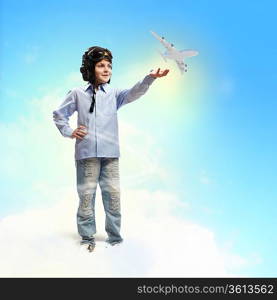 Image of little boy in pilots helmet playing with toy airplane against clouds background