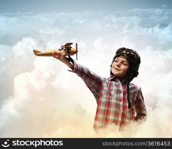 Image of little boy in pilots helmet playing with toy airplane against clouds background