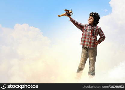 Image of little boy in pilots helmet playing with toy airplane against clouds background