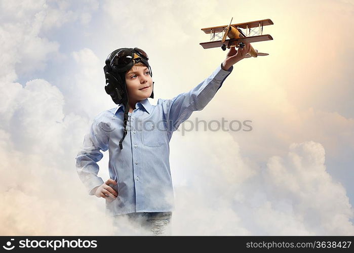 Image of little boy in pilots helmet playing with toy airplane against clouds background