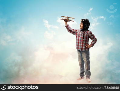 Image of little boy in pilots helmet playing with toy airplane against clouds background